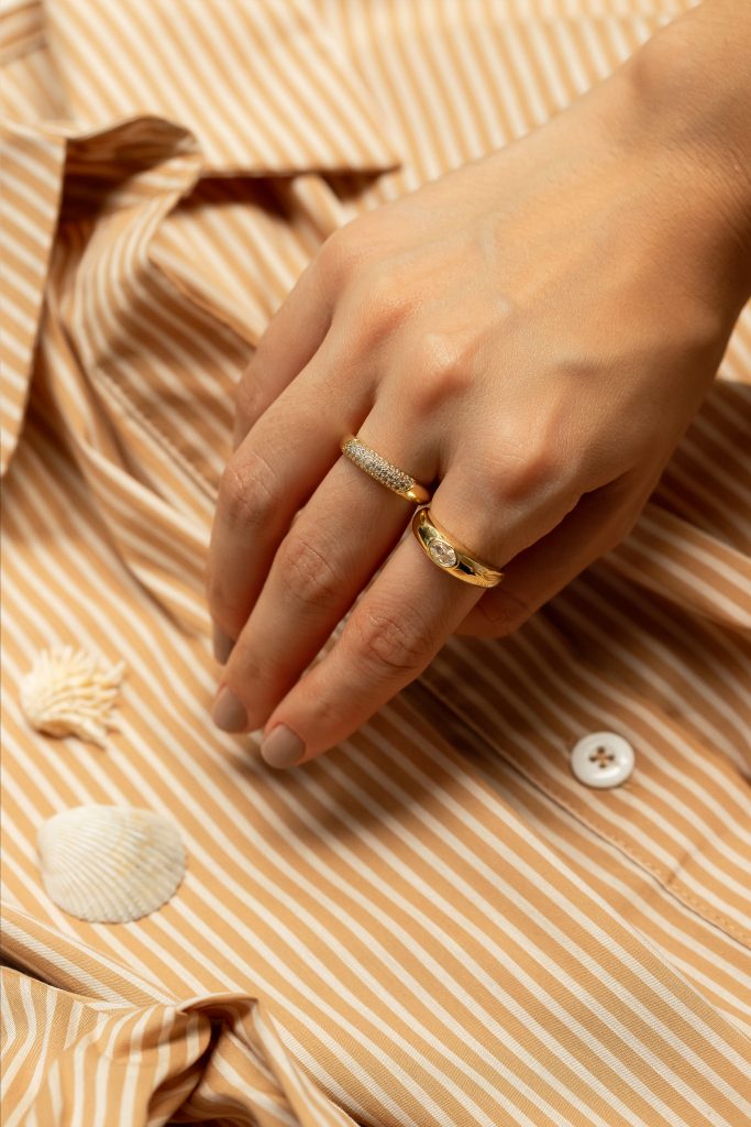 Mão de modelo portando dois anéis no banho de ouro. Ao fundo, uma camisa em tom alaranjado com listras brancas e duas conchas deixam a imagem com um ar típico do verão.