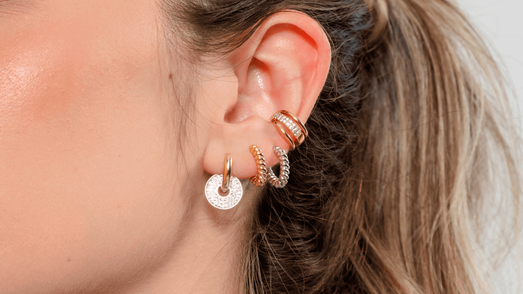 Modelo usando um mix de brincos, que é uma mistura de Ouro e Ródio.
Argolinha com medalha cravejada, duo de argolinhas coração e piercing fake.